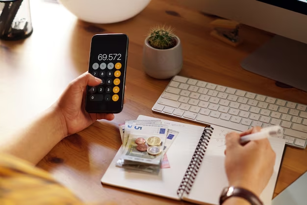 Hand holding a phone with a calculator app, and the other hand holding a pen writing in a notebook, with money placed on top of the notebook.